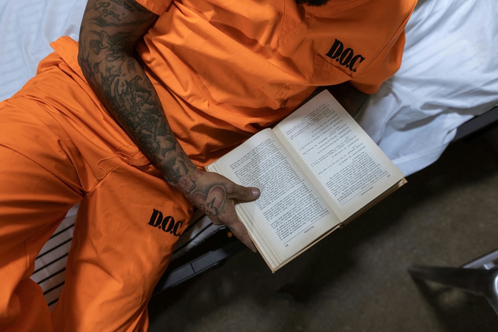 Tattooed inmate reading a book on a bed in a prison cell.